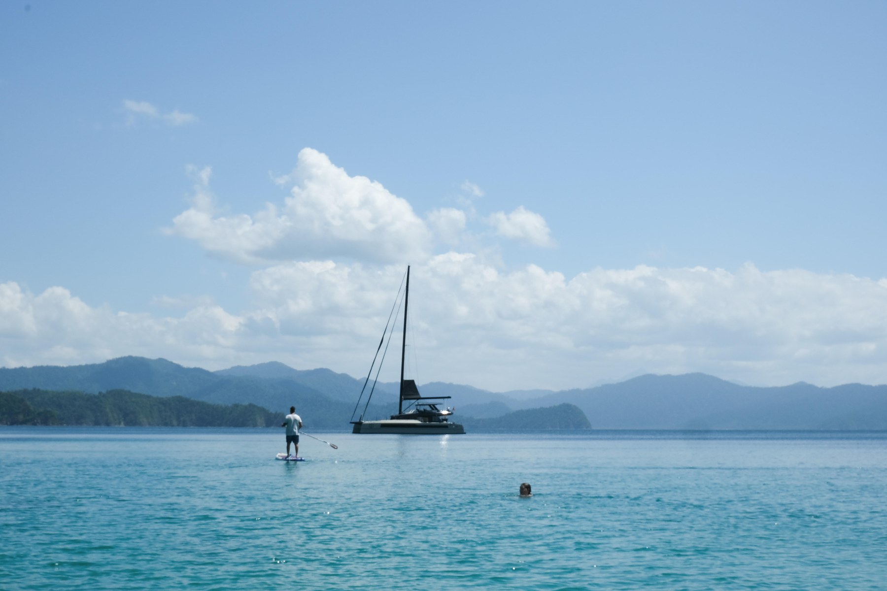 A cruising catamaran at anchor in a calm bay under a big sky, a paddleboarder heading out to her